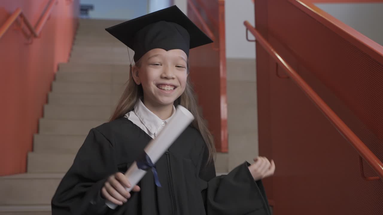 feliz estudiante asiática de preescolar con toga y birrete sosteniendo un diploma y celebrando su graduación