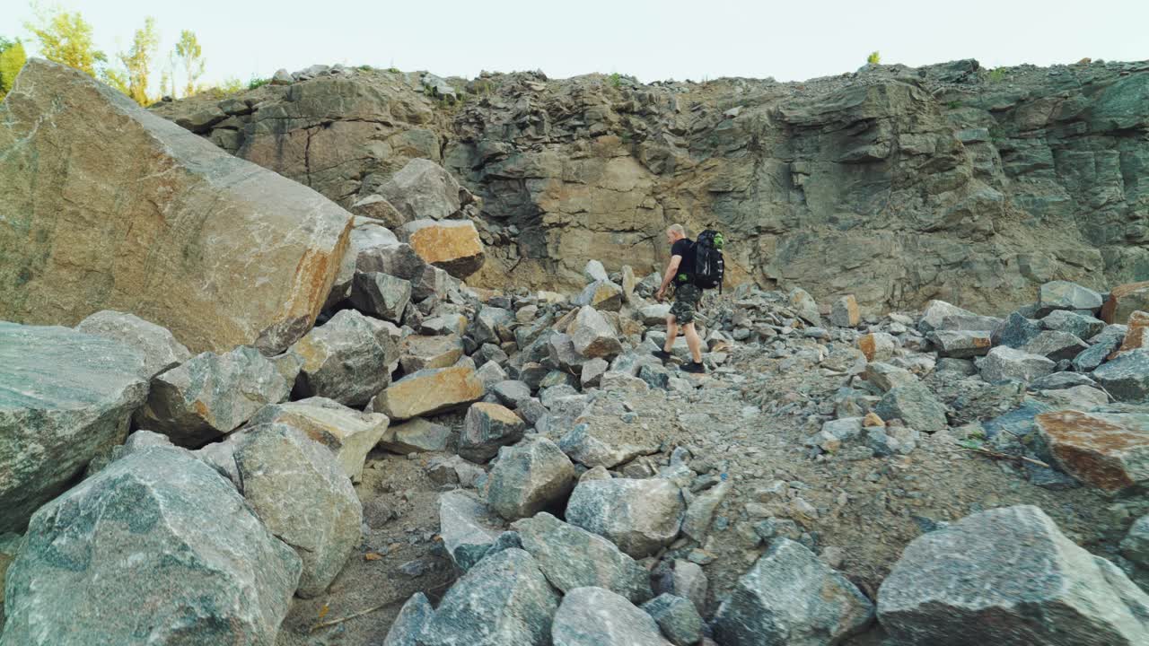 traveler in sportswear with a backpack on his shoulders is going on a pile of stones to a huge stone on the background of rocks in warm summer day