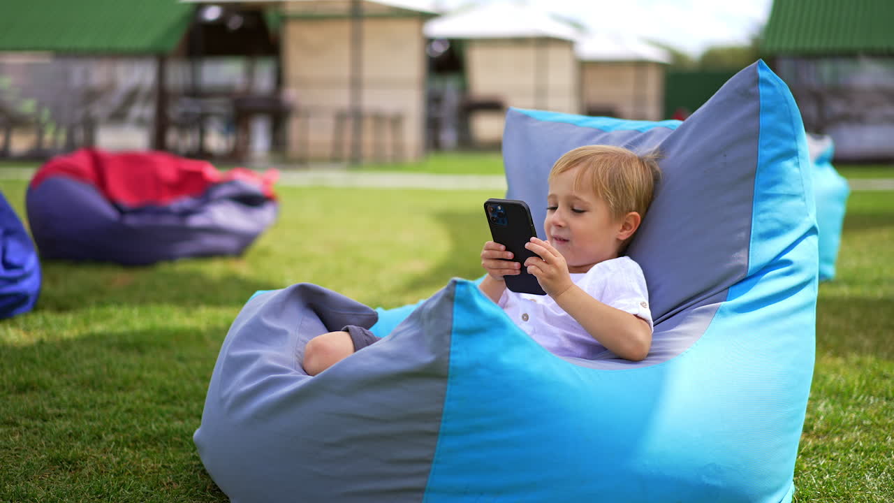 Little boy looking at phone holding it very close to his face. Child in comfortable chair looks aside smiling. Outdoors backdrop in blur.