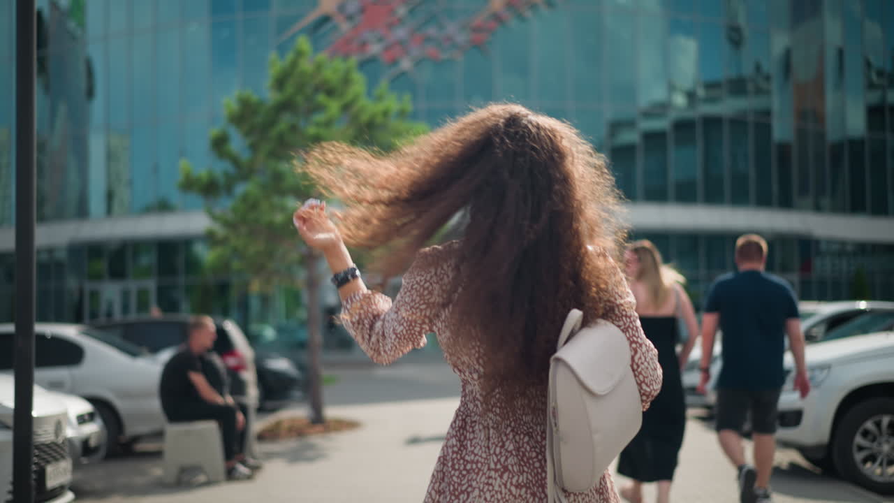 Back view of young adult with curly hair, white bag, wearing hand bangle and smartwatch, walking in urban area with parked cars on both sides and people in background