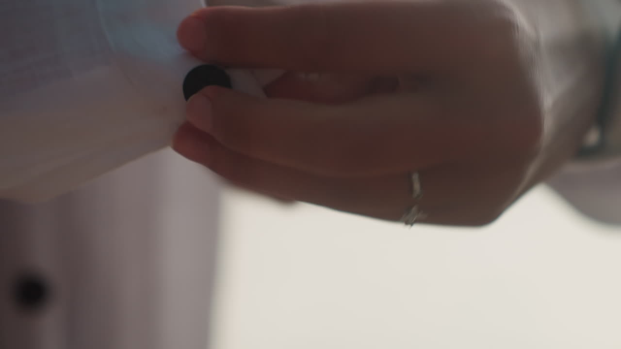Tangan mengancingkan manset kemeja, close-up, wanita mengencangkan kancing hitam kecil pada lengan linen tipis dengan jari-jari lembut, kuku terpoles dan perhiasan halus, bayangan lembut dan cahaya latar, minimalis yang anggun