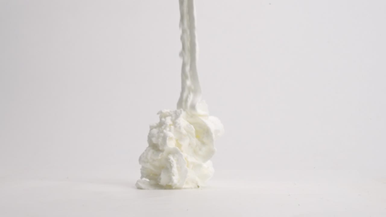 신선하고 푹신한 흰색 휘핑 크림 리본이 흰색 테이블 위에 슬로우 모션으로 쌓여 있습니다.