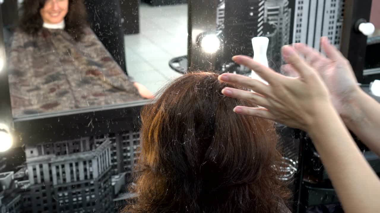 헤어드레스 스타일리스트는 미용실에서 거울 앞에 앉아 머리에 바니쉬를 뿌리는 머리 스타일링 소녀입니다.