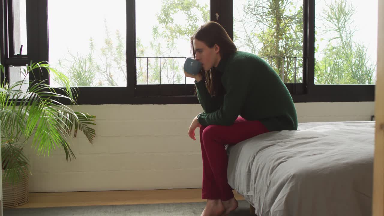 침대에 앉아 커피를 마시는 백인 비 바이너리 트랜스젠더 여성