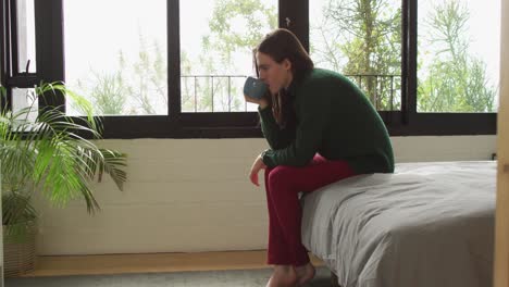 caucasian non-binary transgender woman sitting on bed and drinking coffee