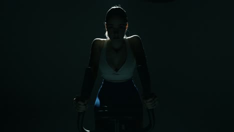 wide shot of a fit woman cycling in a dark gym, camera moving centrally toward her with a slight side slide as cinematic backlight highlights her shoulders and head, capturing power and rhythm