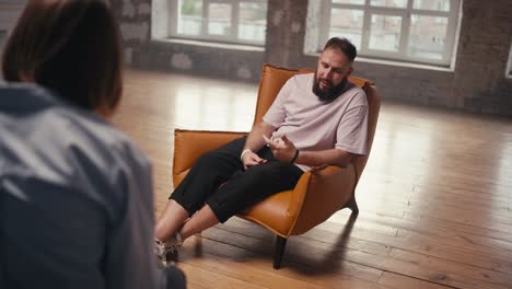 A-man-with-a-beard-in-a-light-pink-brown-chair-talks-about-his-problem-to-a-psychologist-at-group-therapy-in-a-brick-building