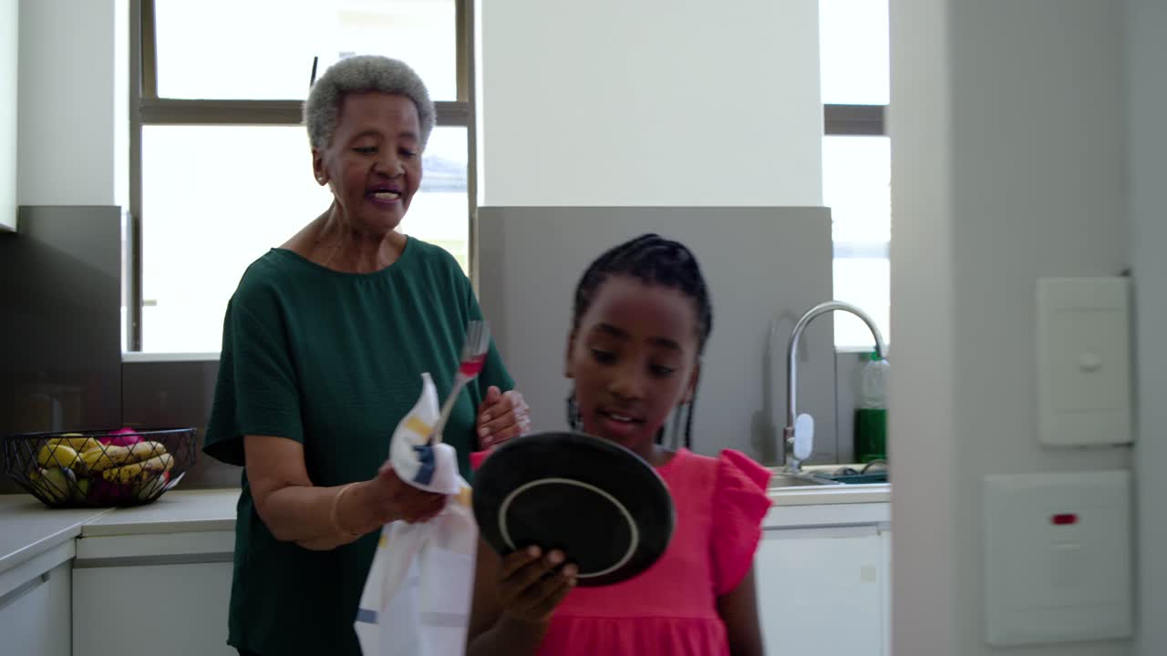 Senior woman holding towel offering plate urging Youth girl checking rim for chips at kitchen sink