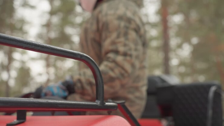 Il pilota di ATV si prepara sul cofano rosso con casco e giacca mimetica, controllando specchio e portapacchi, poi sale sul manubrio. Le scene mostrano i guanti, la presa e un primo piano della saldatura del telaio sullo sfondo di una pineta, in un'atmosfera tesa.