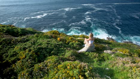 young woman sitting on the rock in the hills with wildflowers and admiring the blue ocean