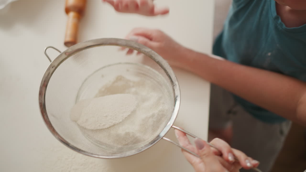 overhead view of child pressing flour through sieve into glass bowl while adult supports from side on white countertop with baking tools like rolling pin visible