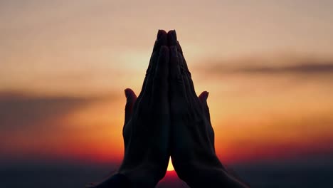 silhouette of a person praying with hands raised as the sun sets, casting warm hues of orange and red in the sky