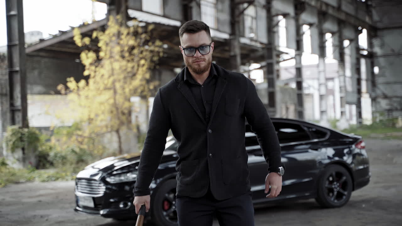 Strong man with a bat in abandoned place. Stylish man in glasses and black wearing moving forward on expensive car background inside the old empty plant.