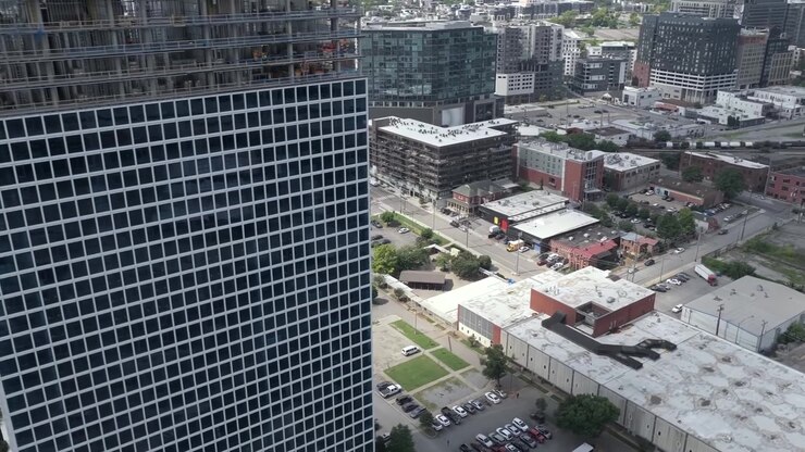 Edifício moderno de arranha-céus passando por expansão vertical em um distrito urbano. Os andares inferiores concluídos apresentam uma fachada de grade geométrica com colunas de concreto aparente, vista aérea orbital