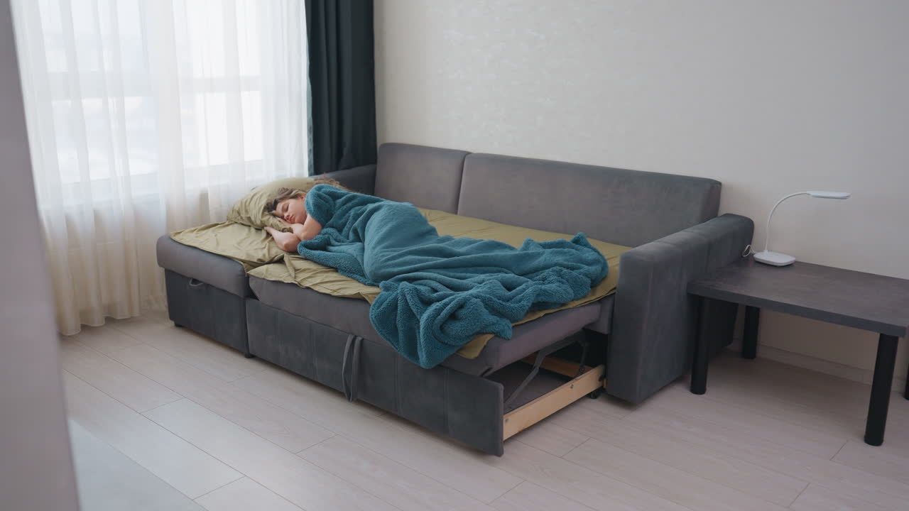 Woman Nurse Resting On Sofa After Long Night Shift, Curled Under Teal Blanket In Bright Apartment Living Room With Sheer Curtains And Side Table Candid Quiet Recovery Moment Conveying Fatigue