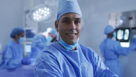 Portrait-of-mixed-race-male-surgeon-standing-in-operating-theatre-smiling-to-camera