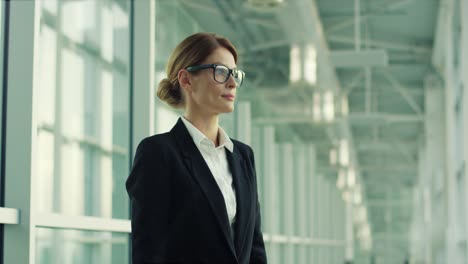 Portrait-Of-The-Beautiful-Businesswoman-In-Glasses-Standing-In-The-Big-Hall-Like-Of-Airport,-Turning-To-The-Camera,-Smiling-And-Crossing-Her-Hands-In-Front-Of-Her