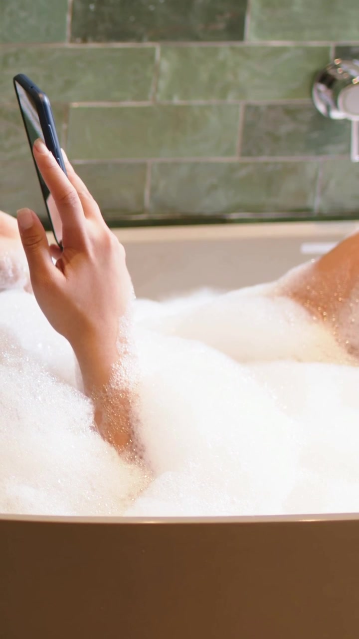 Vertical video: Rising right arm from foam, woman tapping and swiping phone in bathtub, green tiles