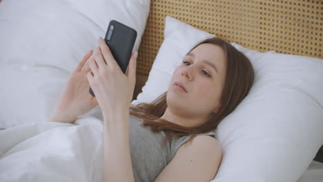 woman in morning sitting in bedroom leaning against the soft leather back of bed. write messages with your smartphone use apps to information