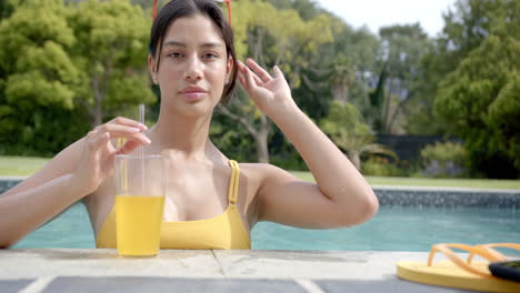 Retrato-De-Una-Feliz-Adolescente-Birracial-Parada-En-Una-Soleada-Piscina-Con-Jugo,-Cámara-Lenta