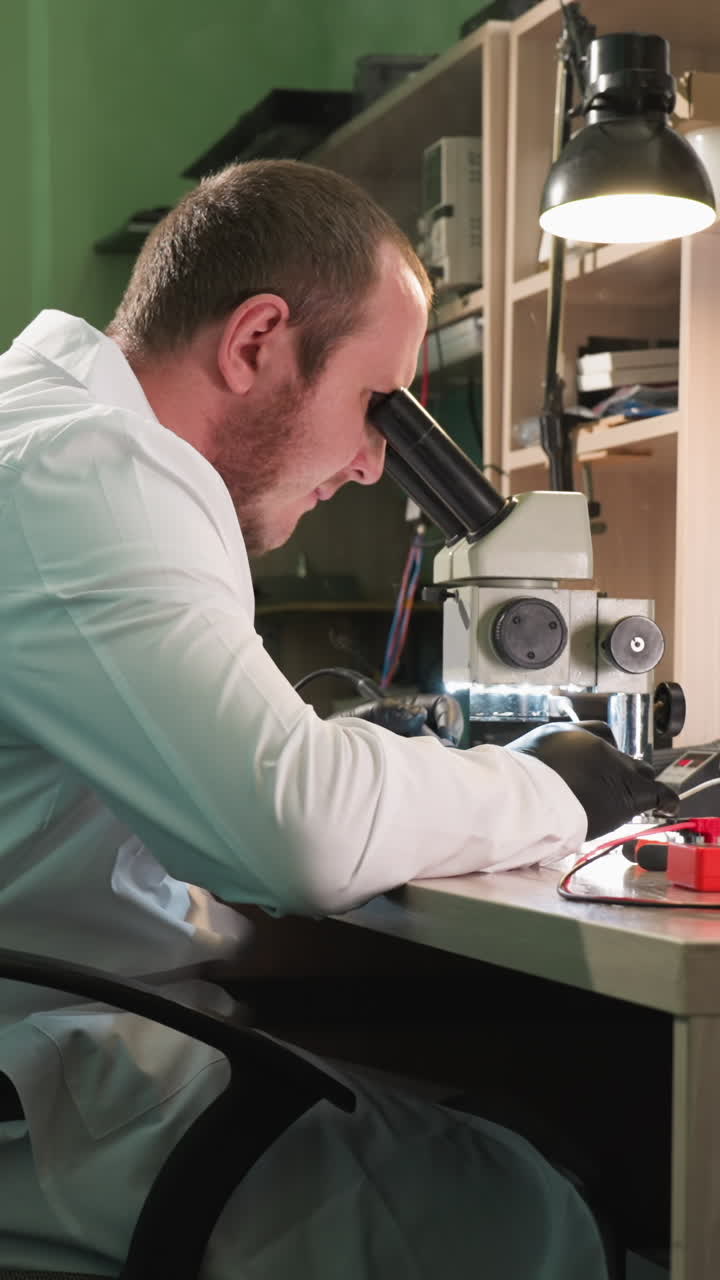 A close-up view of a technician in a white lab coat working intently on a circuit board under a microscope in a well-equipped tech workspace, with a screen on the desk