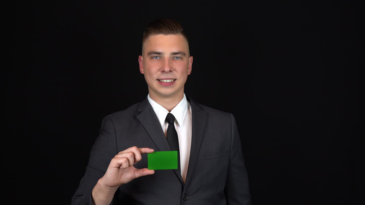 Un giovane uomo d'affari in abito tiene una carta bancaria in mano. Sfondo nero isolato. Carta verde per chromakey.
