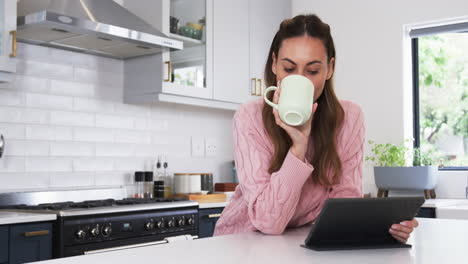 woman in cozy sweater drinking coffee and using tablet in modern kitchen, copy space