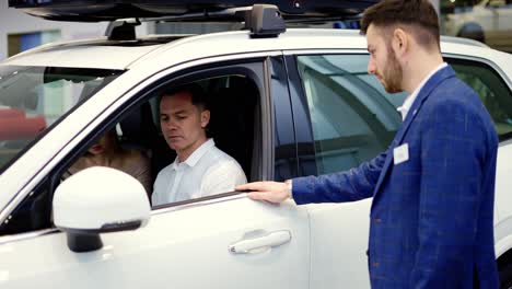 una hermosa pareja de jóvenes se sientan en el automóvil y lo prueban, el gerente de ventas del agente de automóviles ayuda a los clientes