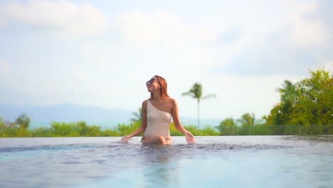 Fröhliche-Frau-In-One-Shoulder-Monokini-Und-Sonnenbrille,-Die-Wasser-Mit-Den-Händen-Spritzt,-Während-Sie-Am-Rand-Eines-Exotischen-Resort-Pools-Mit-Füßen-Im-Wasser-Sitzt,-Vordere-Zeitlupe