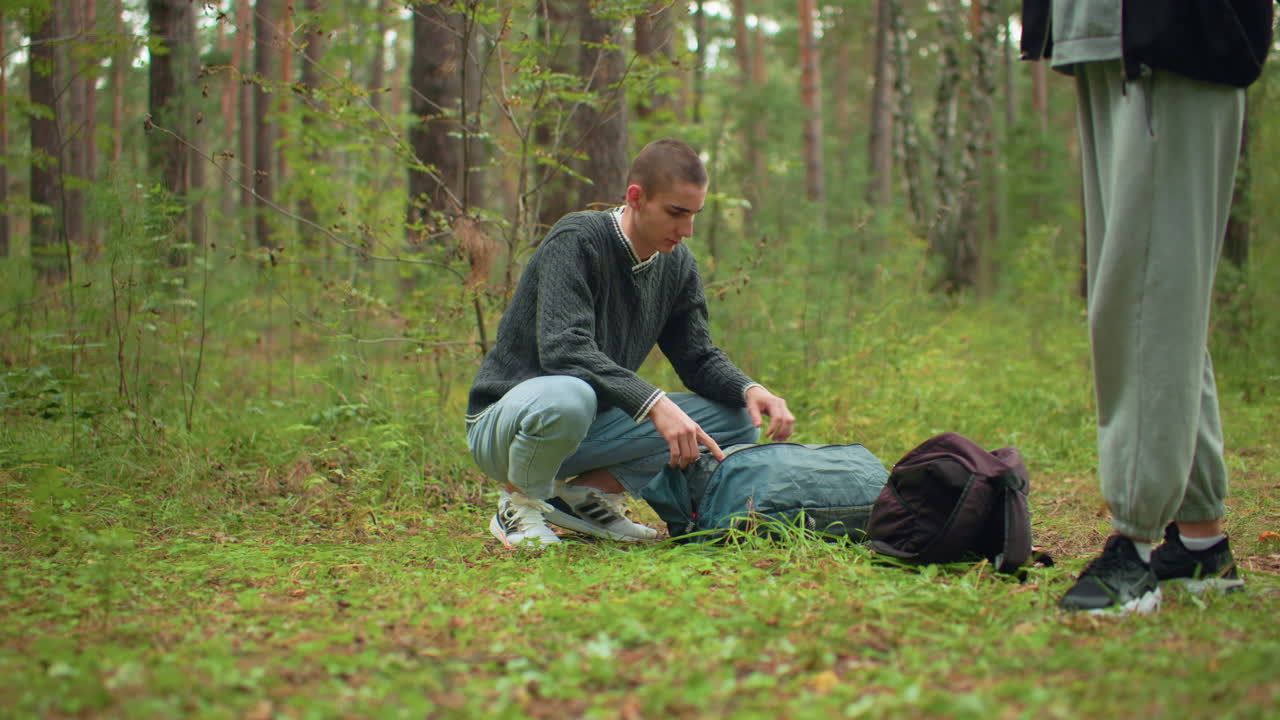 young man crouching in forest unpacking green tent from backpack while woman in casual wear stands nearby stretching arms, surrounded by tall trees and grassy woodland