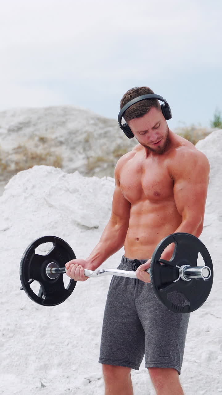 Athletic man with a naked torso in headphones does exercises for the biceps barbell. Fitness in nature. Vertical video