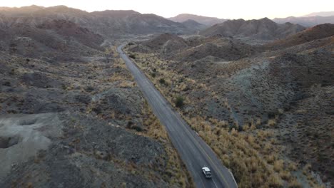 Aerial-drone-following-shot-over-a-white-car-moving-along-a-winding-RCD-road-through-mountainous-terrain-in-Balochistan-during-evening-time