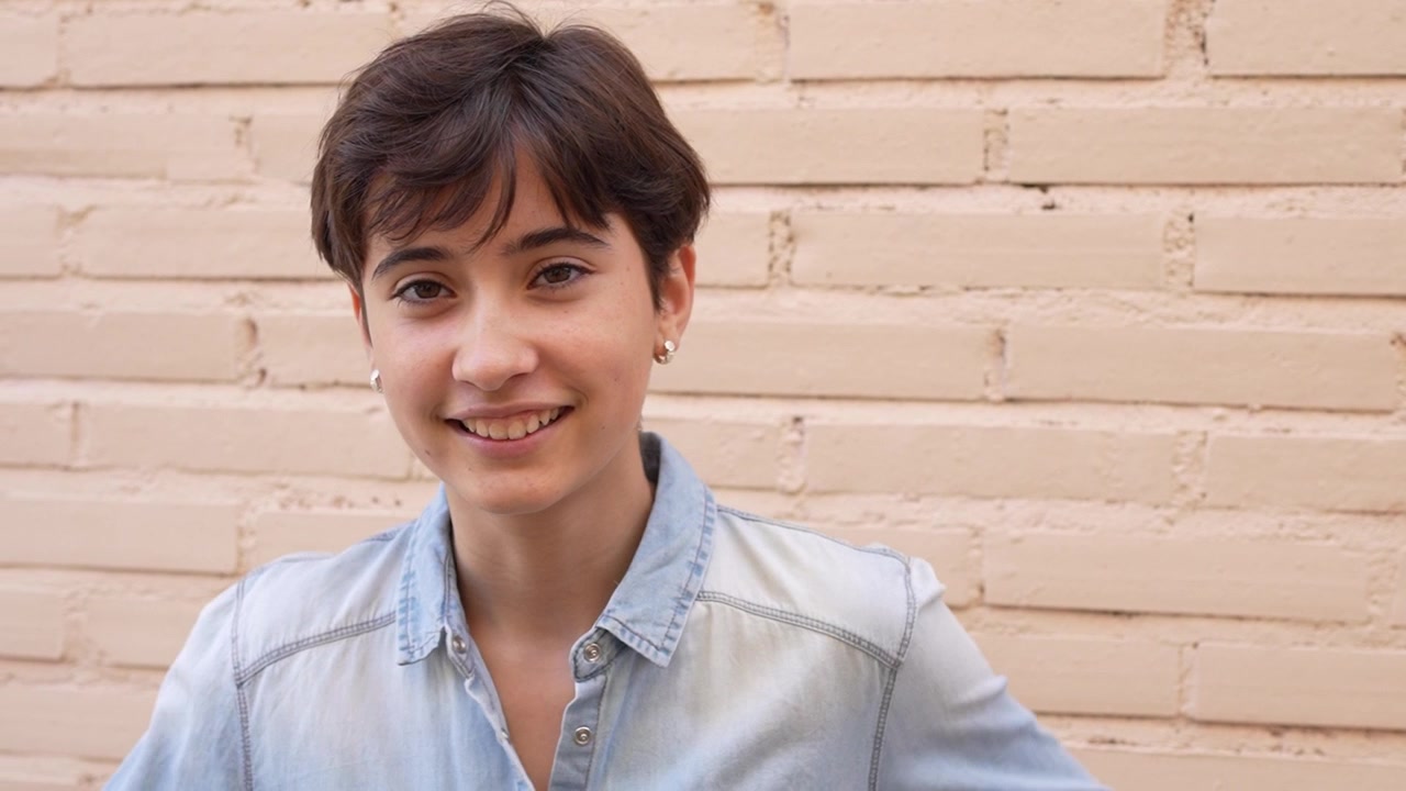 Una giovane persona sorridente che indossa una camicia di jeans casual sta in piedi contro un muro di mattoni rustico