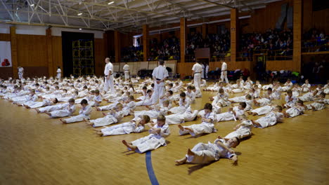 large group of little athletes doing crunches lying on the wooden floor in gym. team of coaches watch the trainees. audience at backdrop.