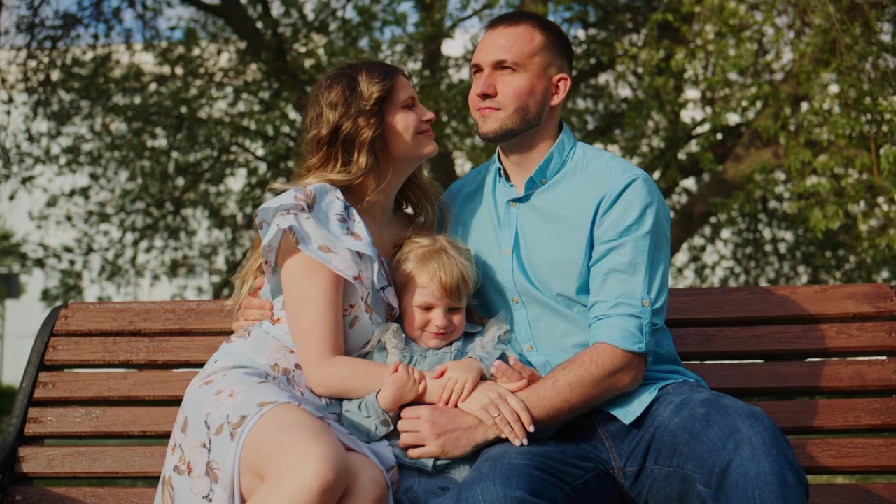 Una famiglia di tre persone siede su una panchina del parco. La madre indossa un vestito floreale, il padre una camicia blu. Il bambino appoggia la testa sulla spalla della madre. Sorridono e si guardano. La scena trasmette affetto e serenità.