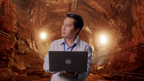 asian male professional worker standing with his laptop in underground mine tunnel, he observes by looking around before he come to concentrating with his laptop