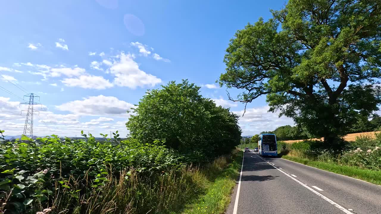 Double-decker bus drives along scenic rural road, sunny summer day, wide landscape, moving camera
