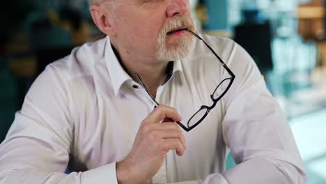 thoughtful businessman wearing white shirt sits at desk. pensive man holds his glasses looking aside. close up.