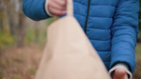 close-up of person in blue sweater flipping brown bag upside down, releasing dry leaves onto the ground in outdoor setting surrounded by autumn foliage and natural environment