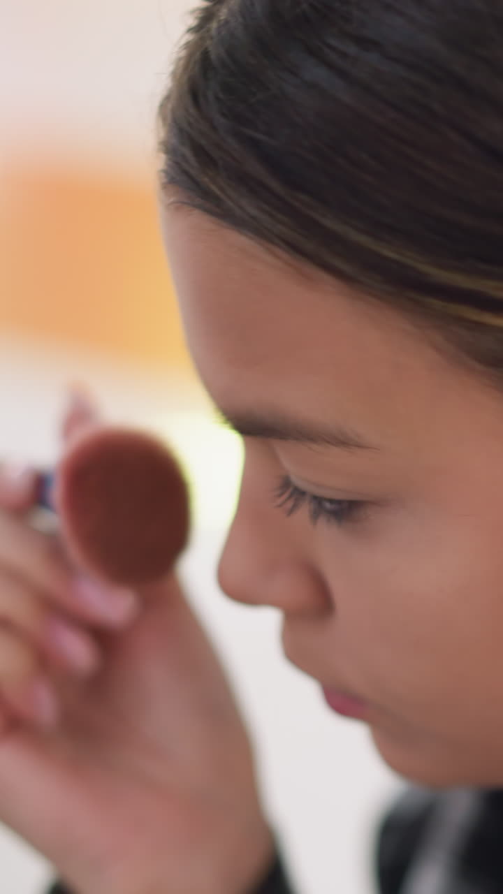 Adolescente practicando maquillaje en el tocador, aplicando polvos y perfeccionando el contorno de la barbilla con un pincel firme. La iluminación suave e íntima y el reflejo en un pequeño espejo capturan la rutina paciente, concentrada y juvenil.