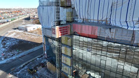 cinematic aerial shot following elevator lift carrying equipment at multi storey building construction site with workers working on floors and revealing cityscape of denver in background