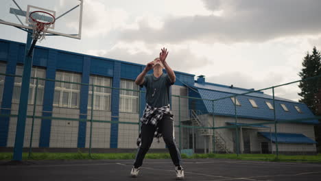 athlete throws volleyball up, jumps, and hits it with basketball court in background, blue building visible, energetic movement, outdoor sport arena, action-filled moment in daylight