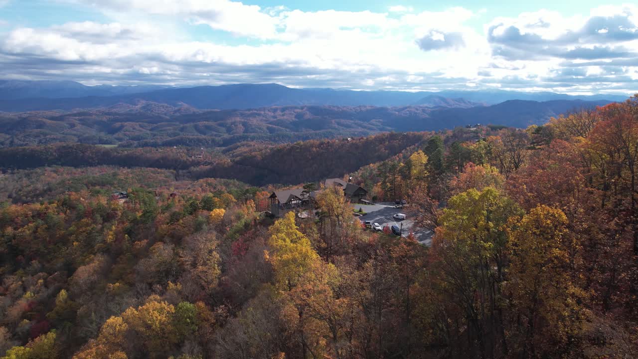 Aerial view zooming in over a forested mountain landscape with vivid autumn foliage and rustic cabins nestled among the trees.