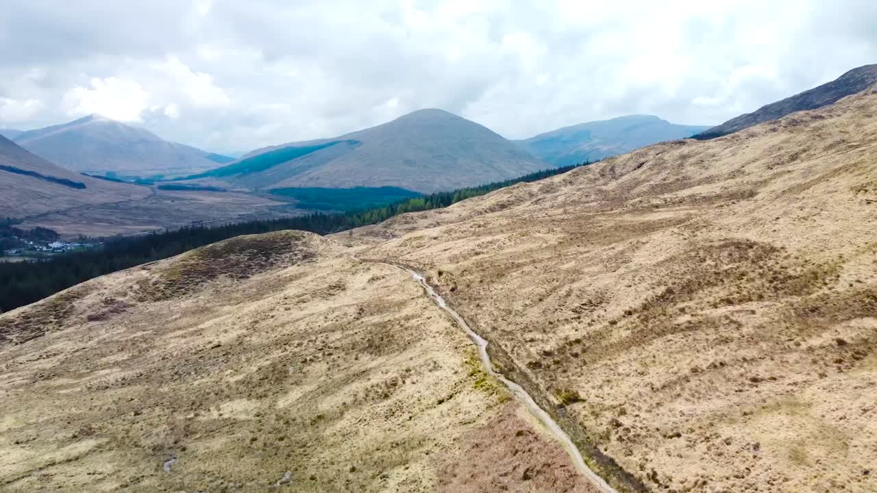 Beautiful aerial drone footage flying forward above grassy yellow scottish highlands landscape where a lonely mud road is leading into the distance during a cloudy day, mountains visible in the back.