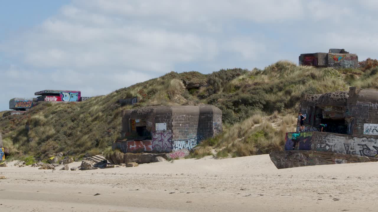 Wide shot of colorful graffiti-covered bunkers on sandy dunes, bright daylight, slow camera pan