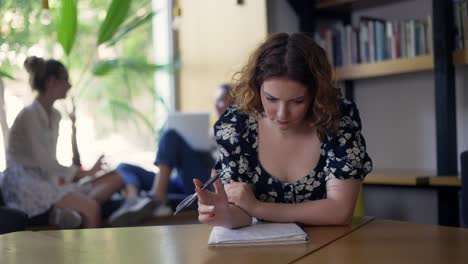 A-young-thoughtful-girl-is-sitting-in-the-library-and-writing-in-a-notebook