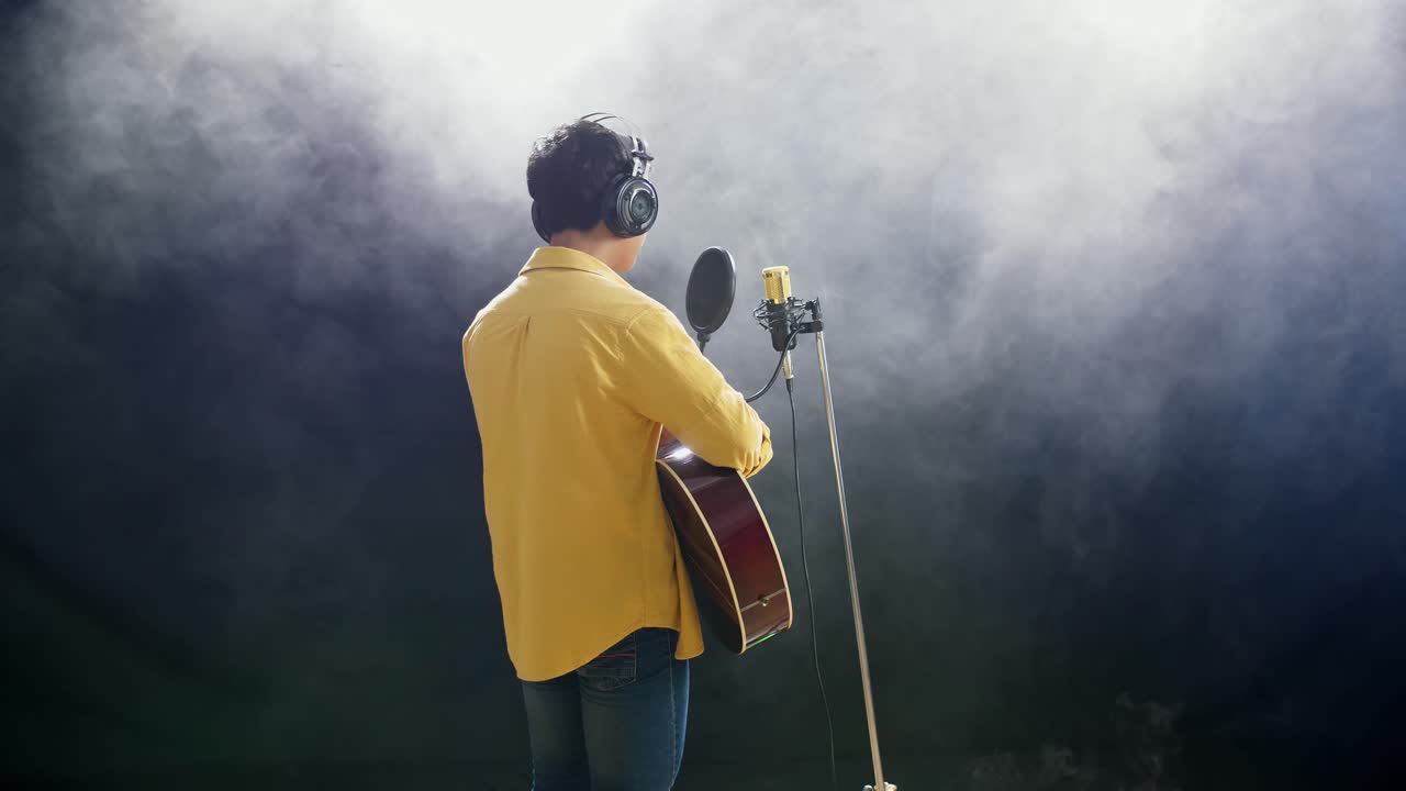 Vista posteriore di un giovane ragazzo con cuffie che suona una chitarra e canta in un microfono a condensatore su sfondo nero con fumo bianco