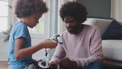 een zwarte vader van middelbare leeftijd op de vloer thuis die zijn tienerzoon helpt een mechanisch speelgoed te bouwen, close-up, zijwaarts