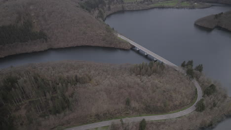 drone disparado en luxemburgo cerca del río lac de la haute-sure en un parque natural öewersauer en un día nublado con coches conduciendo en la carretera cerca del bosque y los árboles y un puente en el registro de fondo