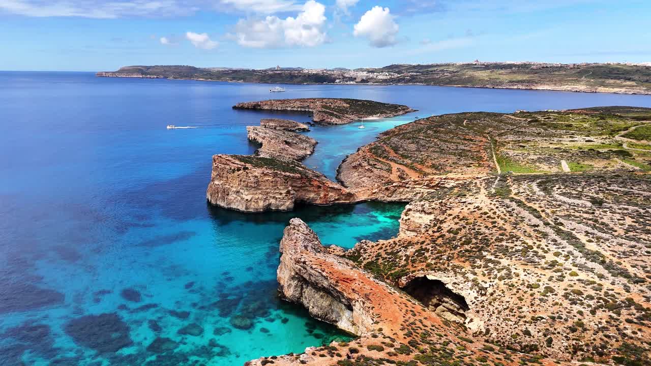 Vista aerea della Laguna Blu a Comino, Malta, che mostra scogliere rocciose aspre, una stretta insenatura turchese e acqua cristallina circondata da un mare blu profondo sotto nuvole sparse.